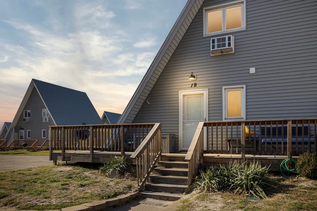 Close-up of the front entrance of a gray A-frame chalet, featuring a wooden deck with railings, steps leading to the front door, and an outdoor grill. An air conditioning unit is visible above a window.