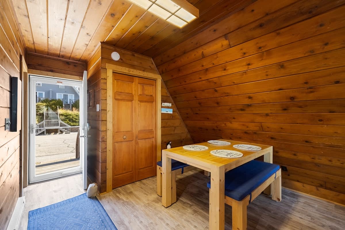 Dining area with a sturdy wooden table and blue cushioned benches, located near the entrance door, featuring wood-paneled walls and a built-in closet with wooden bi-fold doors. The flooring is light wood-look laminate.