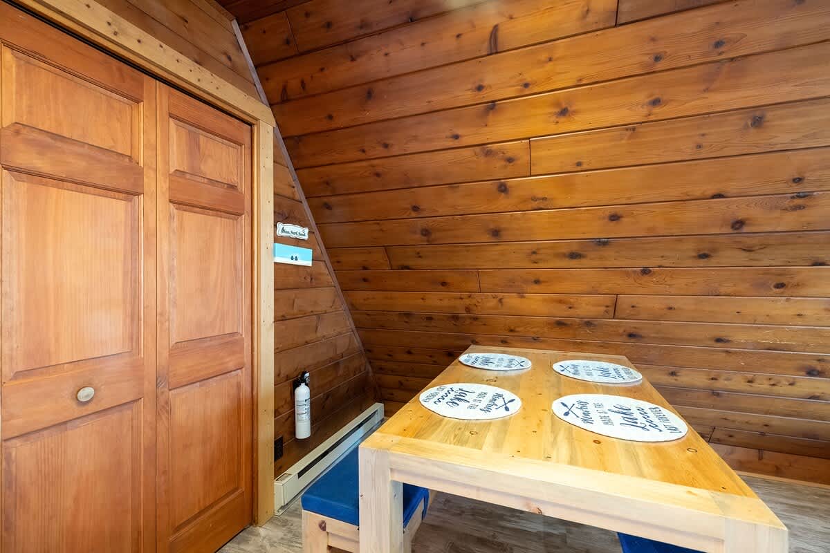 Close-up of the dining area showcasing a light wood table with four place settings and blue benches, positioned against a wood-paneled wall next to a wooden closet with bi-fold doors. The room features a wood-paneled ceiling and light flooring.