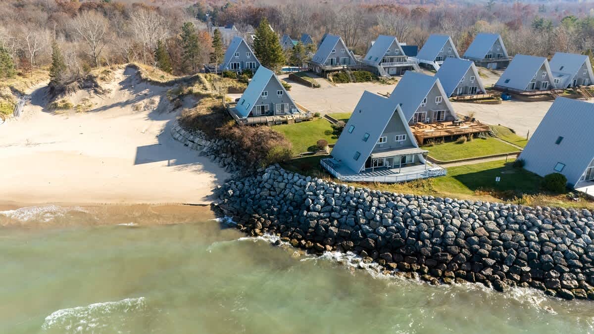 Aerial view showcasing a row of gray A-frame chalets nestled along a sandy beach and rocky shoreline, with a breakwater extending into the water. Lush trees line the background.