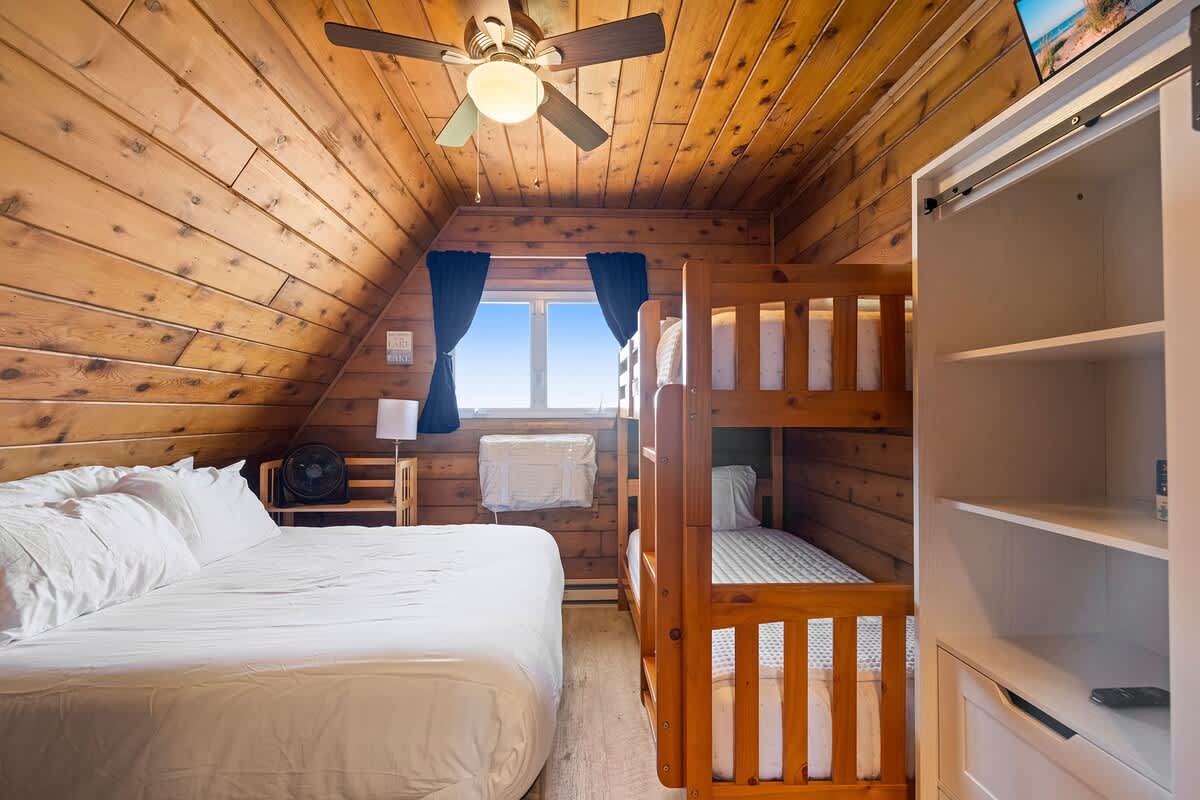 A bright bedroom with sloped wood-paneled walls and ceiling features a queen bed and a wooden bunk bed, with a white open shelving unit for storage. A window with blue curtains offers an outdoor view.