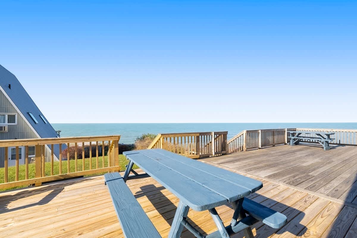 A spacious wooden deck features a blue picnic table in the foreground, with a clear view of the calm blue lake and horizon under a bright sky. A portion of an A-frame chalet is visible on the left.
