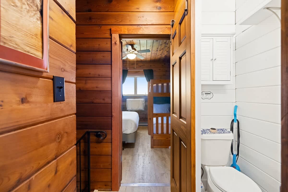 A narrow hallway with wood-paneled walls leads towards a bedroom visible through an open barn door, while a bright white bathroom with a toilet is seen to the right.