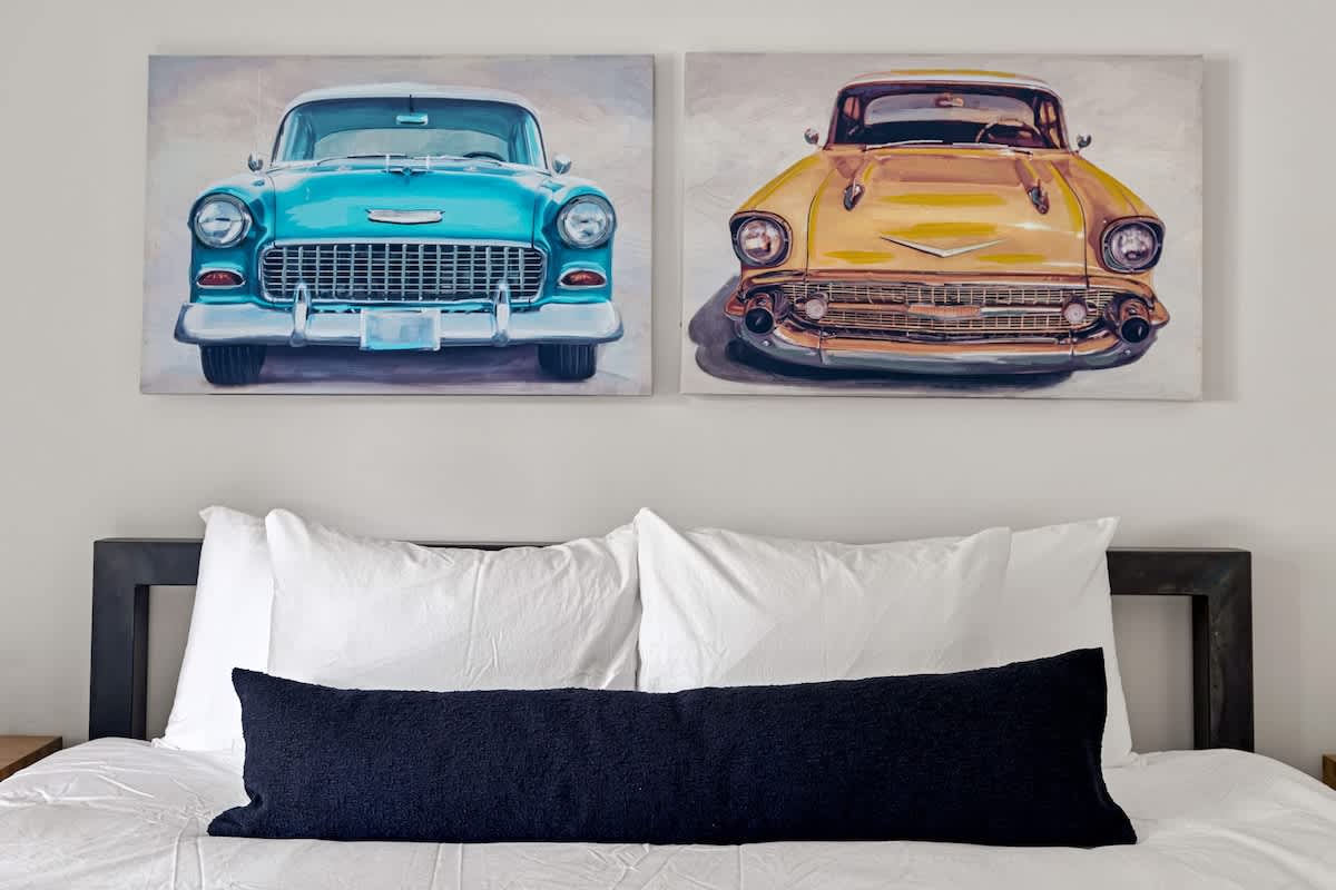 Close-up view of a bed with crisp white bedding and a black bolster pillow, complemented by two vibrant art prints of classic blue and yellow cars hanging above the headboard.