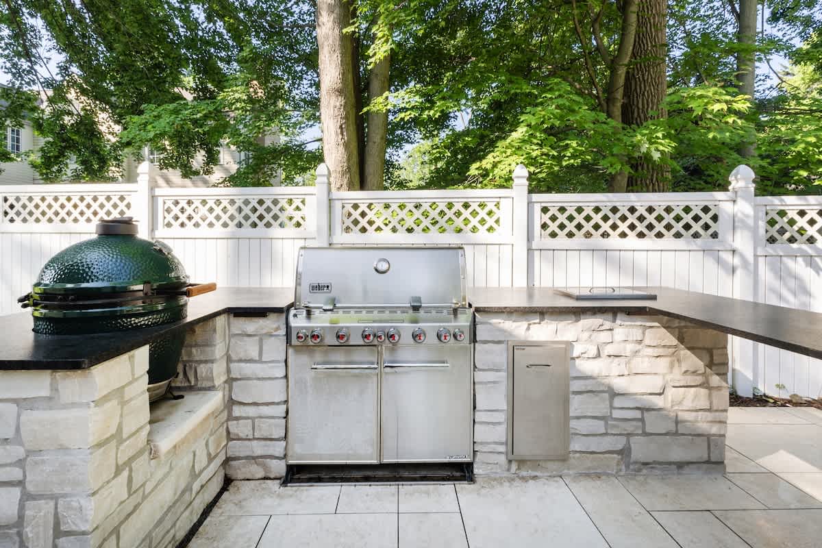 An outdoor kitchen area features a large stainless steel Weber grill and a green Big Green Egg smoker, built into a light stone counter with dark countertops, all against a white lattice fence and lush trees.