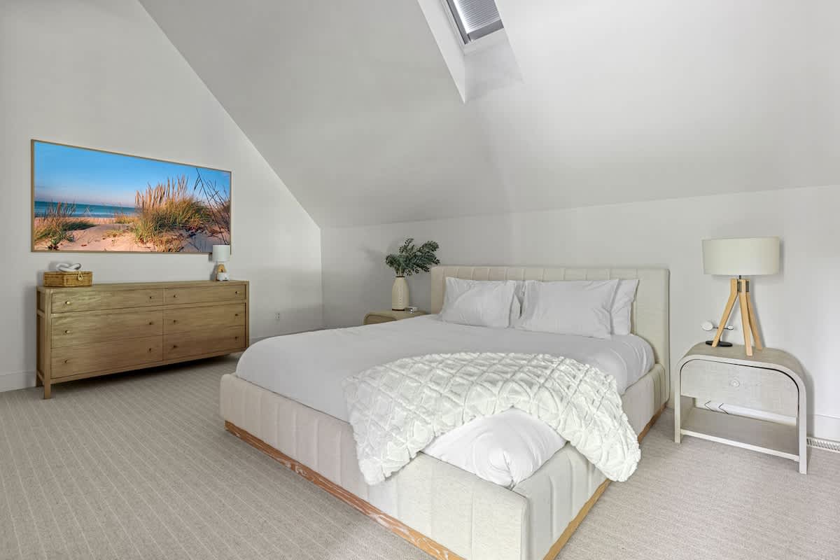Bright bedroom with white walls and a sloped ceiling, featuring a light-colored upholstered king bed, a wooden dresser with a large TV, and a modern nightstand with a lamp.