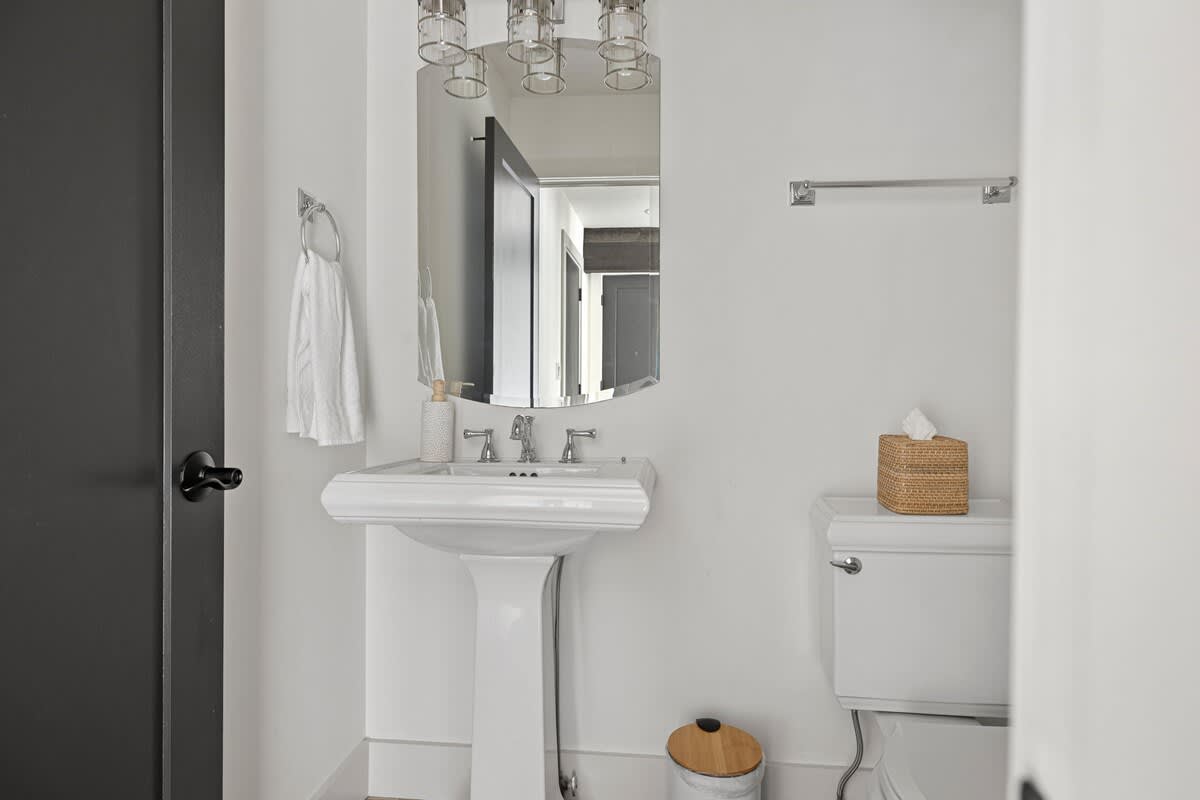 Clean, modern bathroom featuring a white pedestal sink with a rectangular mirror and chrome vanity light, a toilet, and a small woven tissue box on the tank.