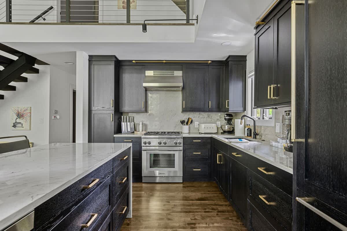 View of the kitchen showing dark cabinetry, white marble countertops, a stainless steel range, and an island with drawers, with a glimpse of a staircase and mezzanine in the background.