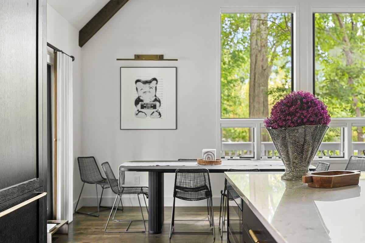Modern dining area with a black rectangular table, wire-frame chairs, and large windows overlooking lush greenery, adjacent to a white kitchen island.