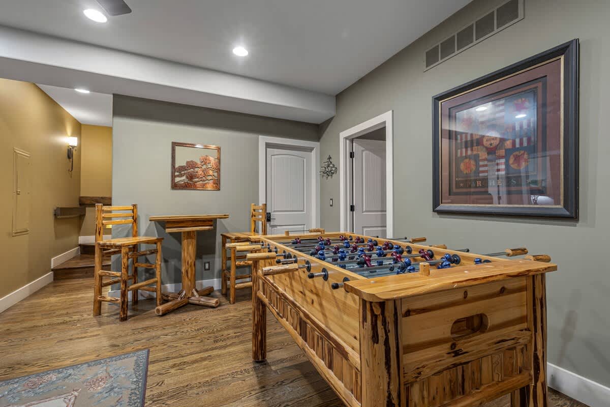 Recreation room with hardwood floors and light green walls, featuring a rustic wooden foosball table and a high-top table with stools, leading to other rooms.