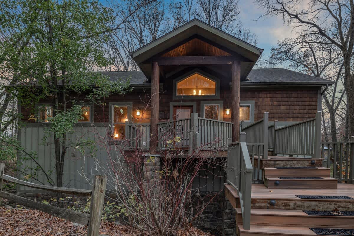 A rustic lodge-style house with dark wood siding and a prominent front porch/balcony, featuring a gabled roof and large windows, nestled among bare trees. A wooden staircase leads up to the main entrance, with stone accents at the base.
