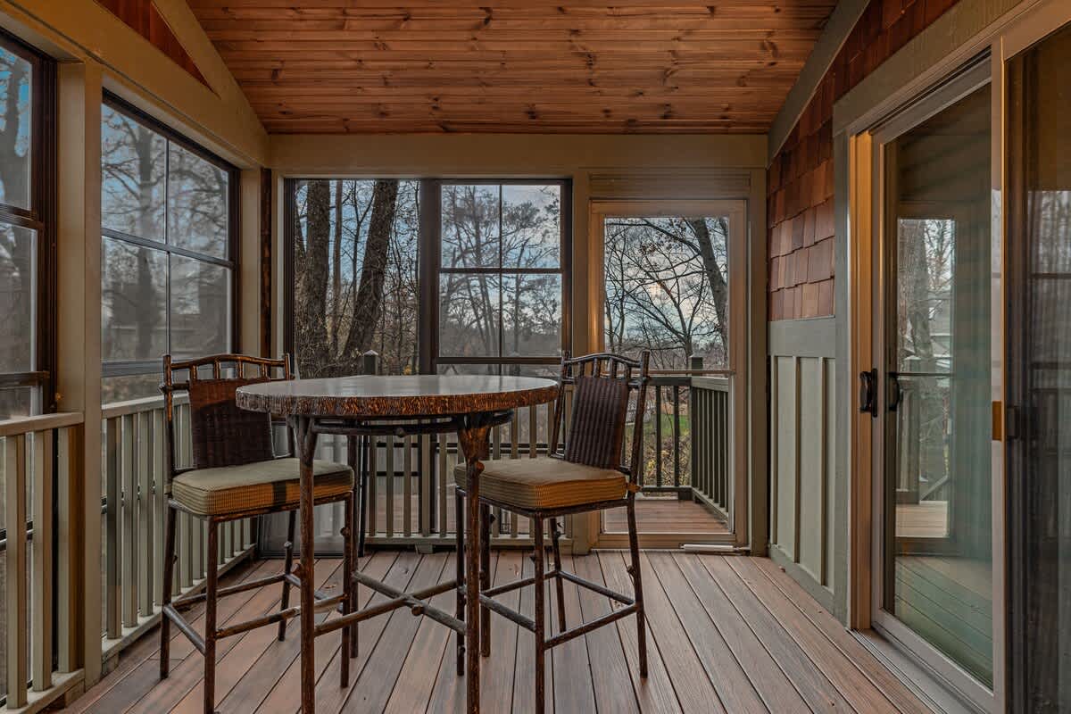 Enclosed screened porch with a small round high-top table and two chairs, offering views of the surrounding trees, with a wooden floor and ceiling.