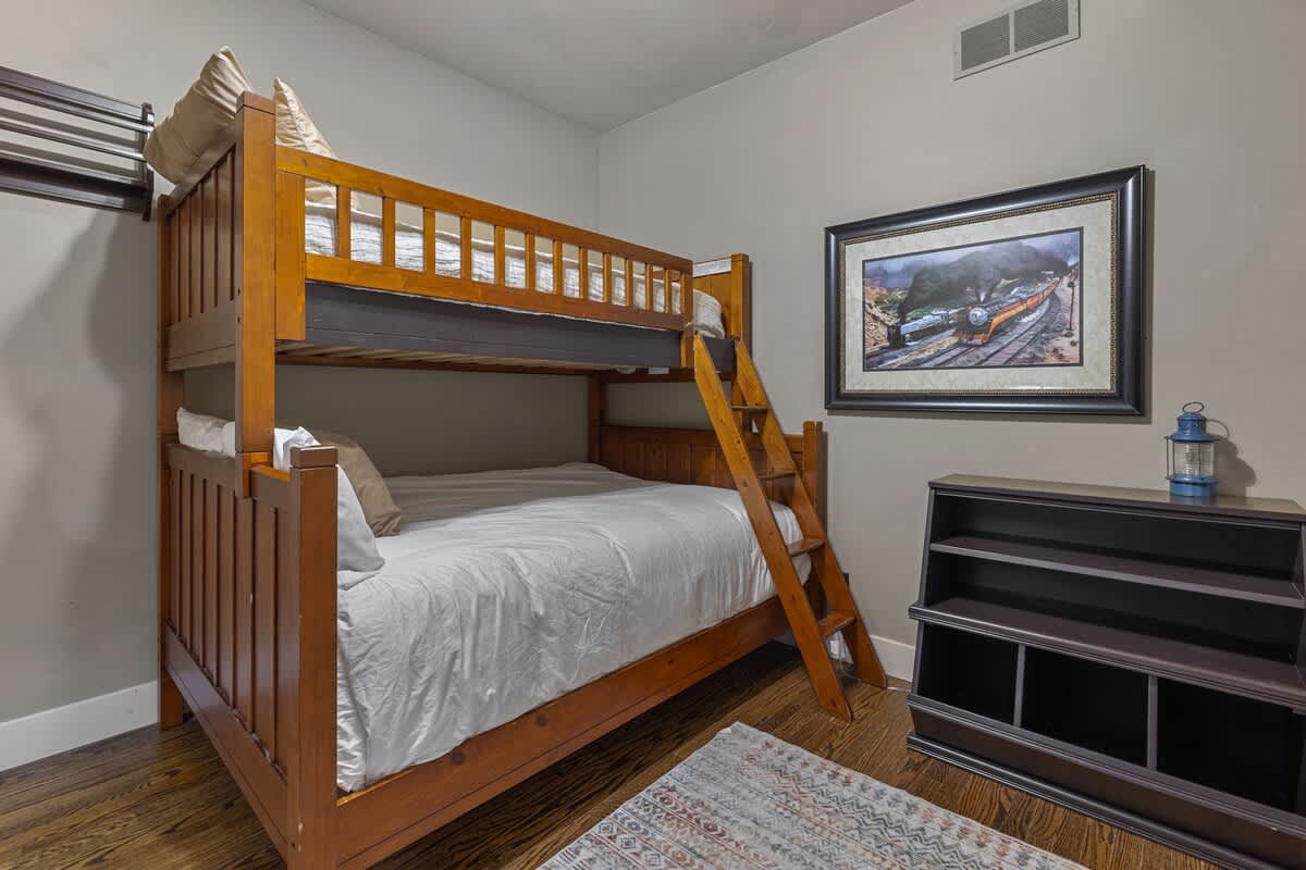 Children's bedroom with a sturdy wooden bunk bed, hardwood floors, a framed train artwork, and a dark wood bookshelf, offering a cozy sleeping arrangement.