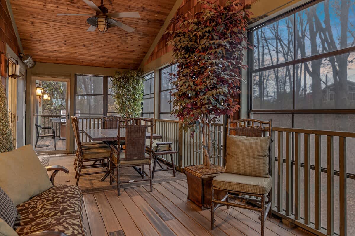 Spacious screened porch with a wood plank ceiling, ceiling fan, and light-colored wood flooring, furnished with a dining table and chairs, and a sofa, offering views of the surrounding trees.