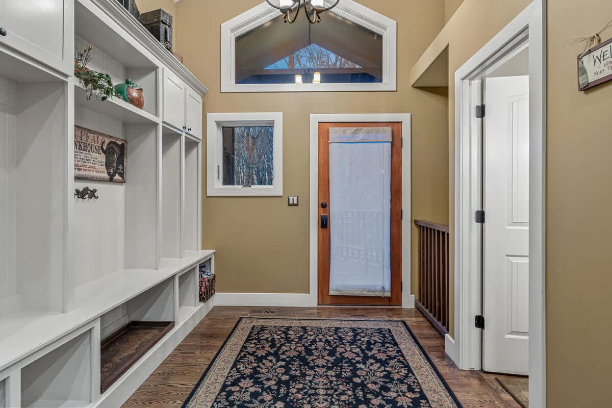 A bright entryway features white built-in cubbies and shelving along one wall, a dark patterned rug on hardwood floors, and a wooden door leading outside. A high arched window above the door lets in natural light.