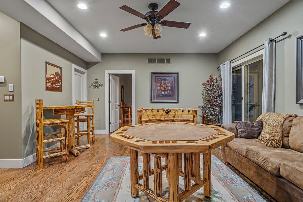 Another view of the game room, highlighting the rustic wooden poker table, a foosball table in the background, and a small high-top table with stools, all on hardwood floors.