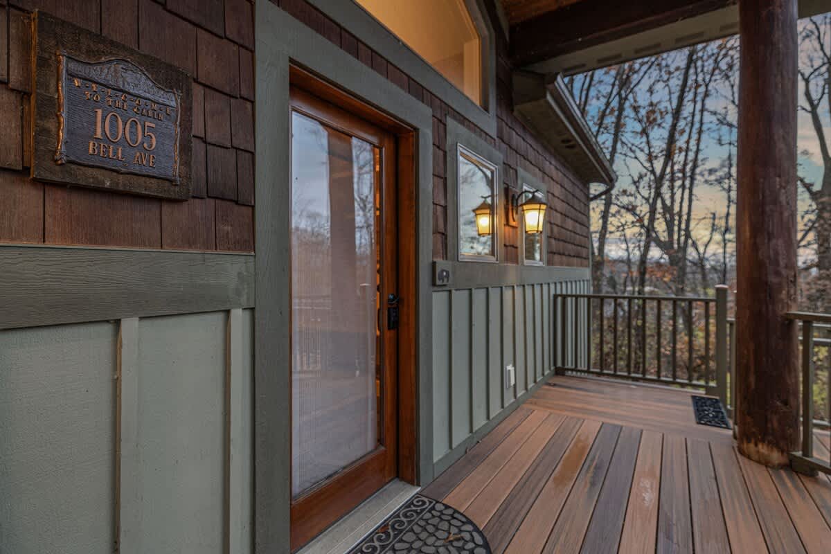 Front entrance of the lodge-style house with a wooden door and sidelights, set on a wooden deck with railings, featuring wall sconces and cedar shake siding.