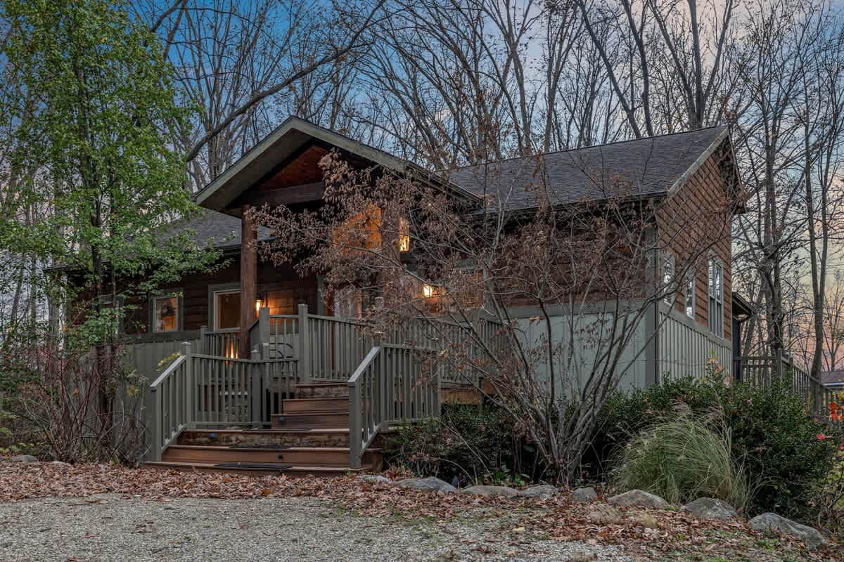 The front exterior of a rustic lodge-style house with dark wood siding and a covered entrance featuring a large wooden deck and stairs. The surrounding area is wooded with bare trees and sparse autumn foliage.