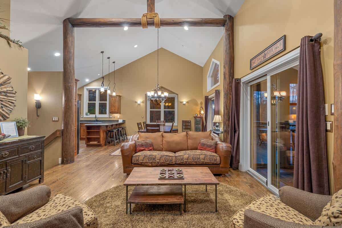 Wide view of the rustic living room with vaulted ceilings, exposed wooden beams, and hardwood floors, showing a leather sofa, armchairs, and a coffee table, with the kitchen and dining area in the background.