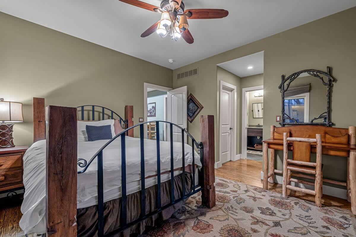 Warm bedroom with a large bed, a rustic wood and metal frame, a patterned rug, and a wooden vanity with a mirror, leading to an en-suite bathroom through an open doorway.
