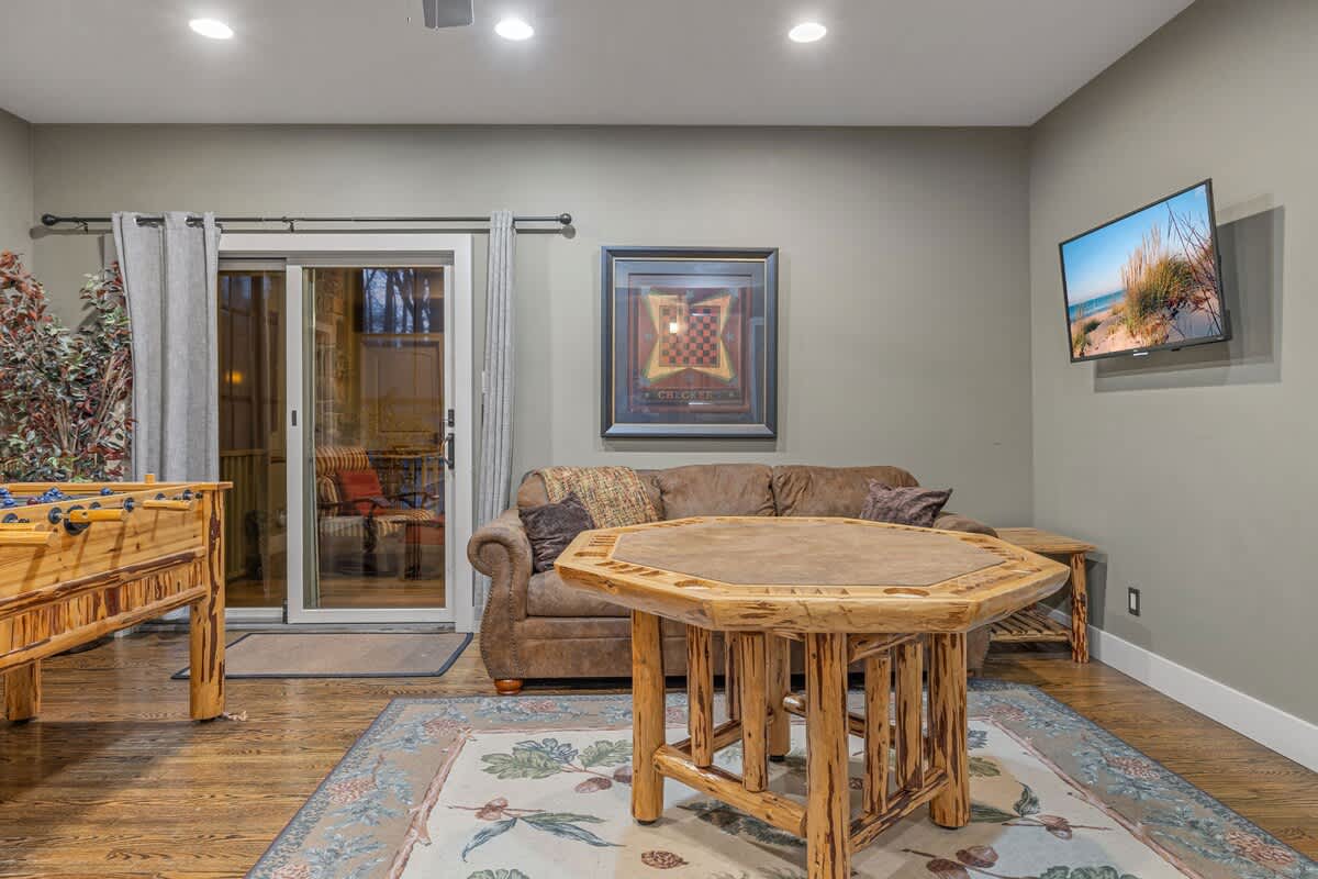 A game room featuring a foosball table, a rustic wooden poker table, a brown leather sofa, and a wall-mounted TV, with a sliding door leading to an outdoor space.