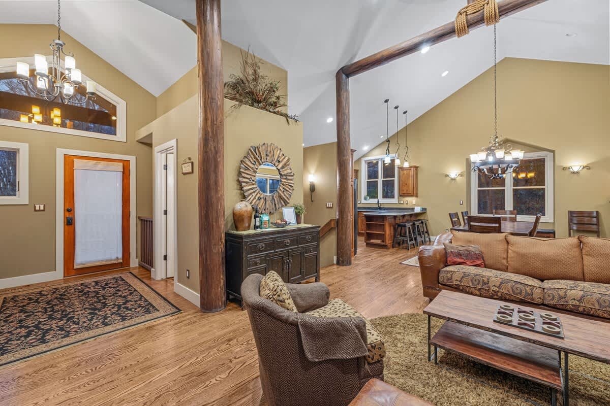 Open-concept living area with vaulted ceilings and exposed wooden beams, featuring hardwood floors, a dark wood console table with a sunburst mirror, and a view towards the kitchen and dining areas.