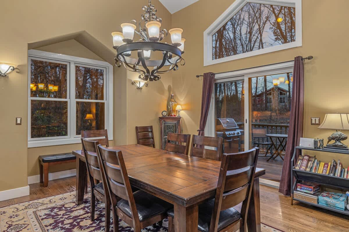 A rustic dining room with a large wooden table and eight chairs, illuminated by a wrought iron chandelier, and featuring large windows overlooking a wooded area.