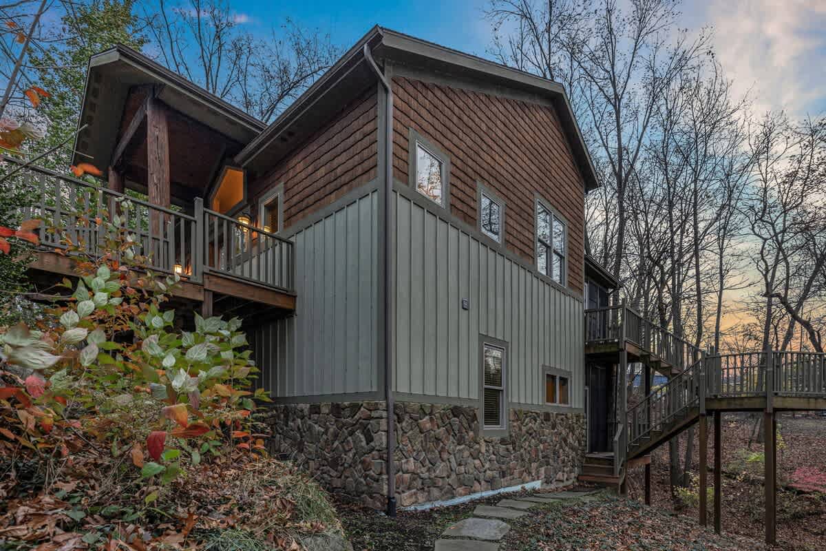 The side exterior of a multi-level lodge-style house, featuring a mix of brown shingle siding, light green vertical siding, and a stone foundation. Wooden decks and stairs lead up to various entrances, surrounded by autumn foliage.