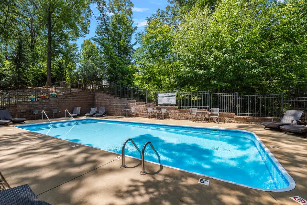 A large rectangular outdoor swimming pool with blue water and metal handrails is surrounded by a concrete deck with lounge chairs. The pool area is fenced and backed by a tiered stone wall and lush trees.