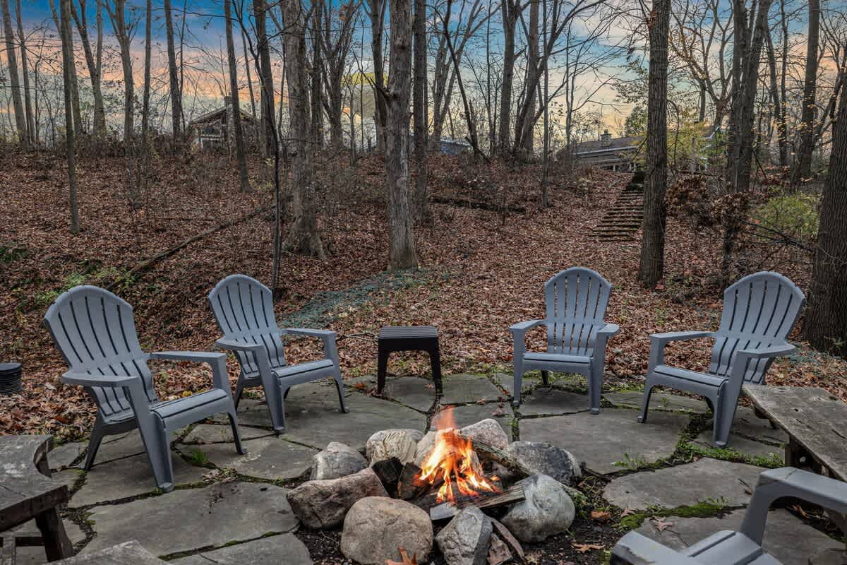 An outdoor fire pit area features a stone-ringed fire pit with a roaring fire, surrounded by four gray Adirondack chairs on a flagstone patio. The setting is a wooded backyard with bare trees and fallen leaves.