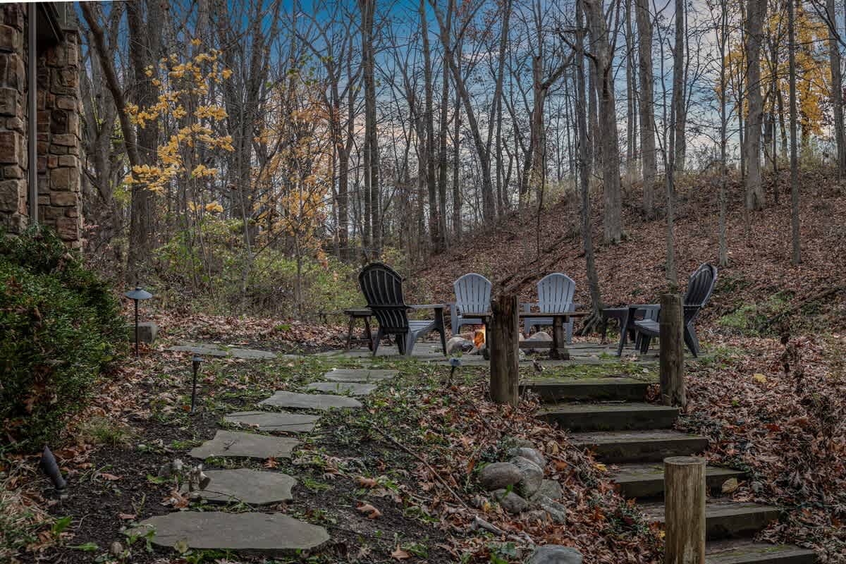 Outdoor fire pit area with a flagstone patio and Adirondack chairs, nestled in a wooded area with fallen leaves and a stone pathway leading up to the house.