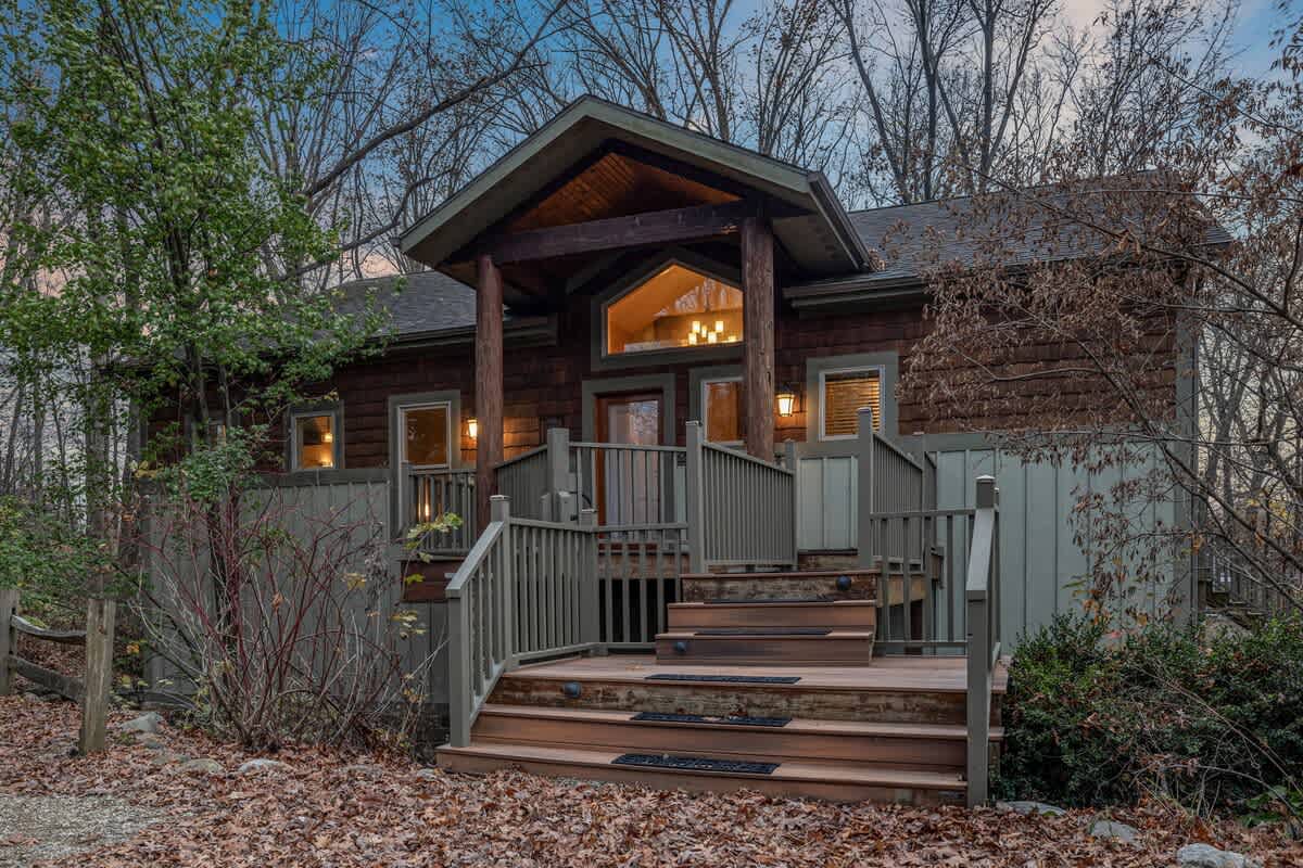 The front exterior of a rustic lodge-style house with dark wood siding and a covered entrance featuring a large wooden deck and stairs. The surrounding area is wooded with bare trees and fallen leaves.