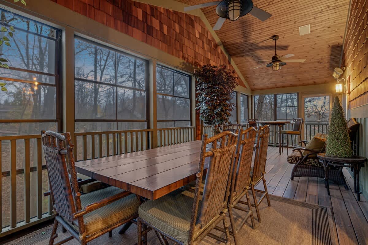 Spacious screened porch featuring a large wooden dining table with multiple chairs, additional seating areas, and ceiling fans, surrounded by windows overlooking nature.