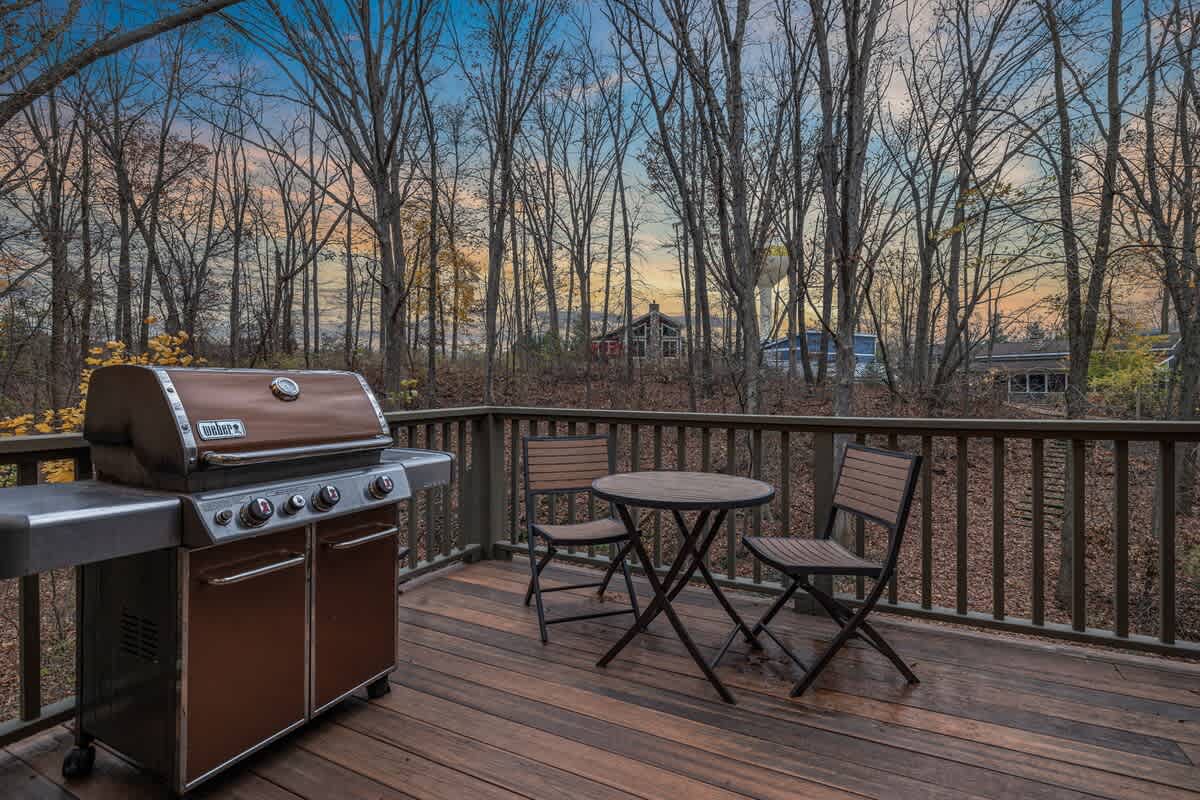 An outdoor wooden deck features a large brown Weber gas grill and a small round bistro table with two chairs, set against a backdrop of bare trees under a colorful sky.