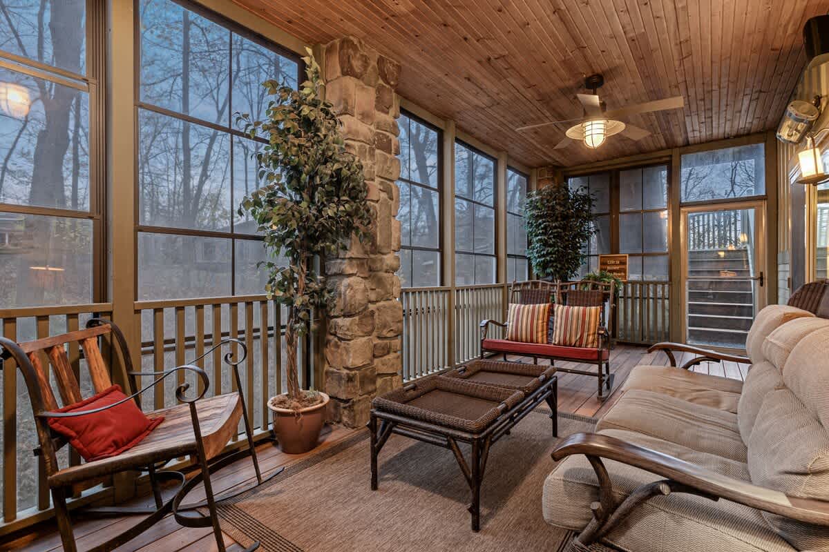 A spacious screened porch with a wooden slatted ceiling and floor, featuring a stone pillar and comfortable outdoor seating. A ceiling fan and wall sconces provide lighting, overlooking a wooded area.
