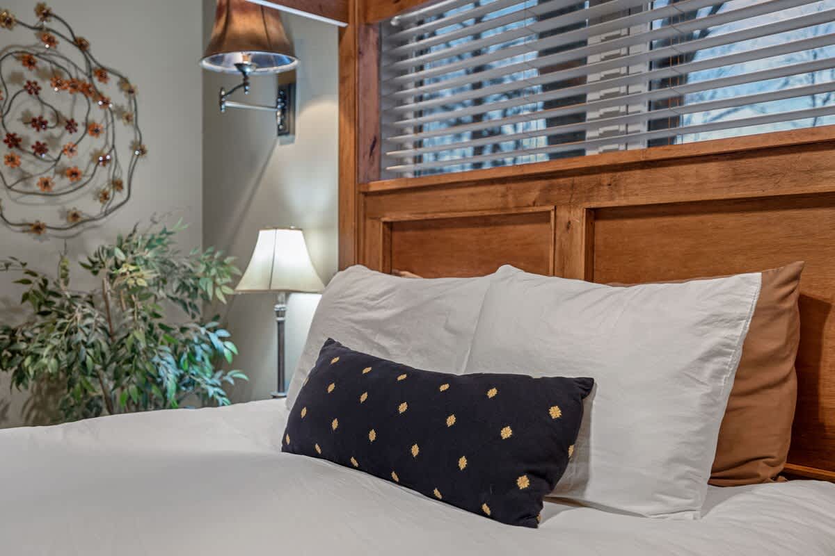 A close-up of the rustic wooden four-poster bed with white bedding and a decorative black and gold pillow, showing a nightstand with a lamp and a plant beside it.