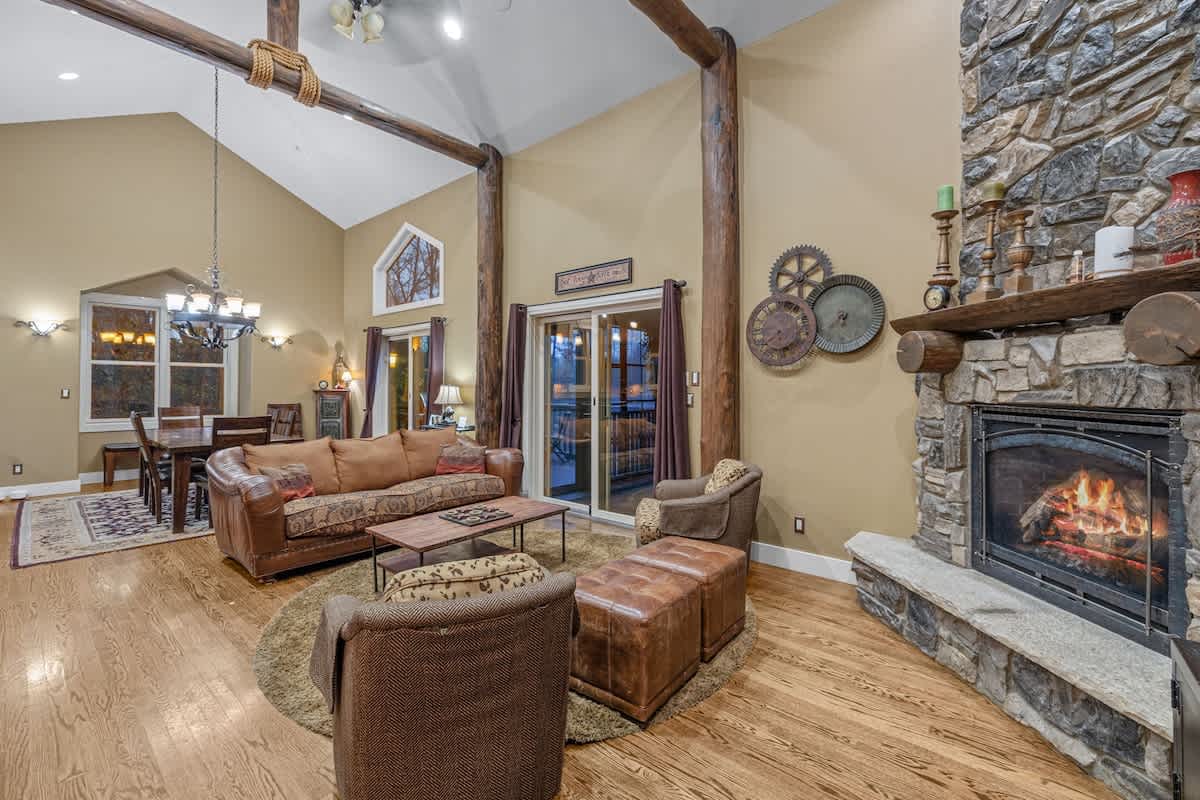 Rustic living room with vaulted ceilings, exposed wooden beams, and hardwood floors, featuring a large stone fireplace, a leather sofa, and two armchairs around a coffee table. A dining area is visible in the background.