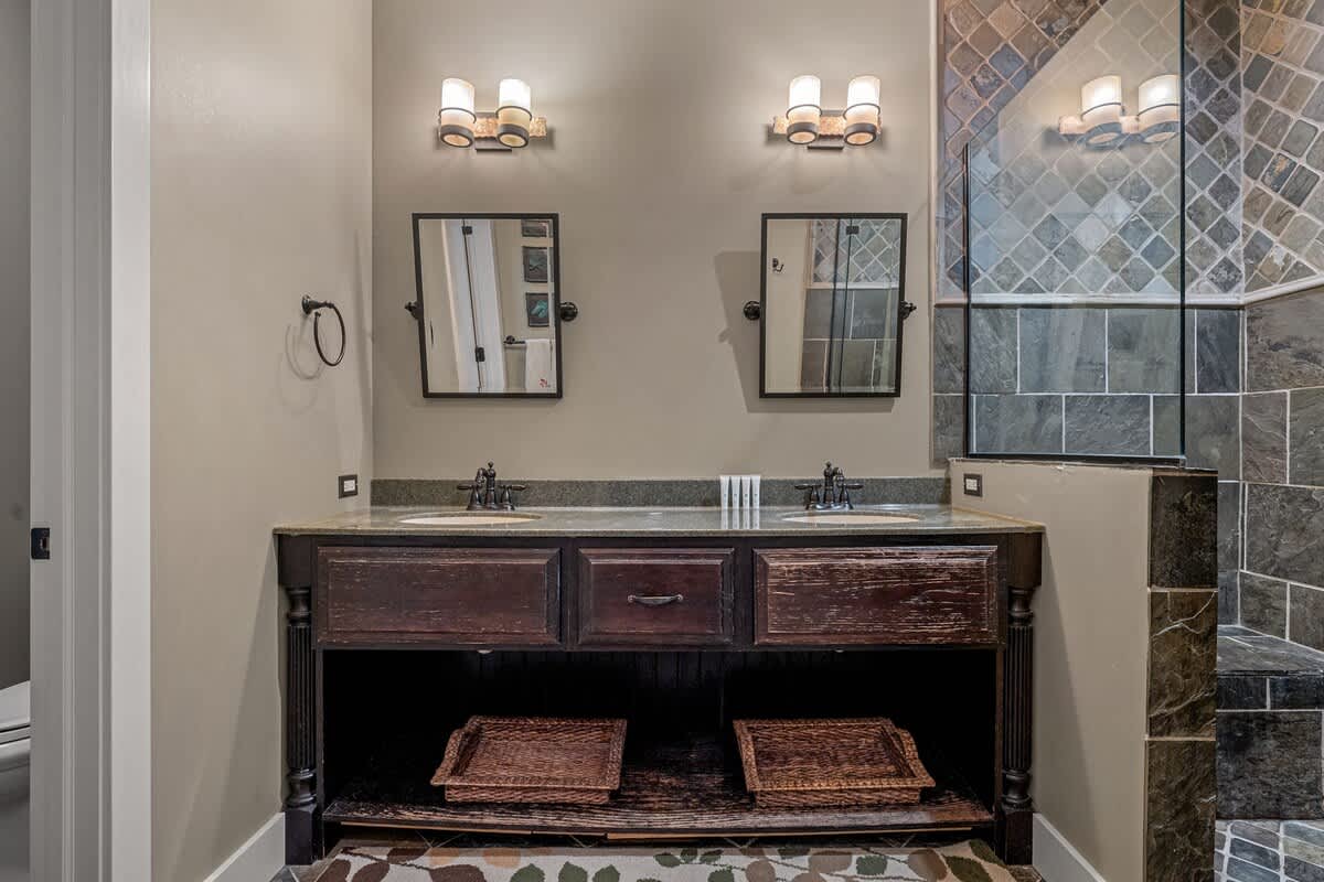 Spacious bathroom featuring a dark wood double vanity with two sinks, framed mirrors, modern wall sconces, and a large walk-in shower with intricate stone tile work.