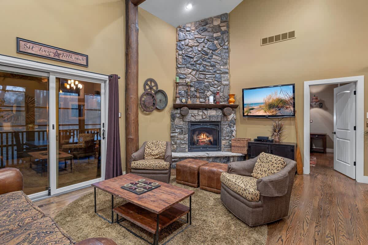 A cozy living room features a tall stone fireplace, two patterned armchairs, a wooden coffee table, and a large sliding glass door leading to an outdoor deck.