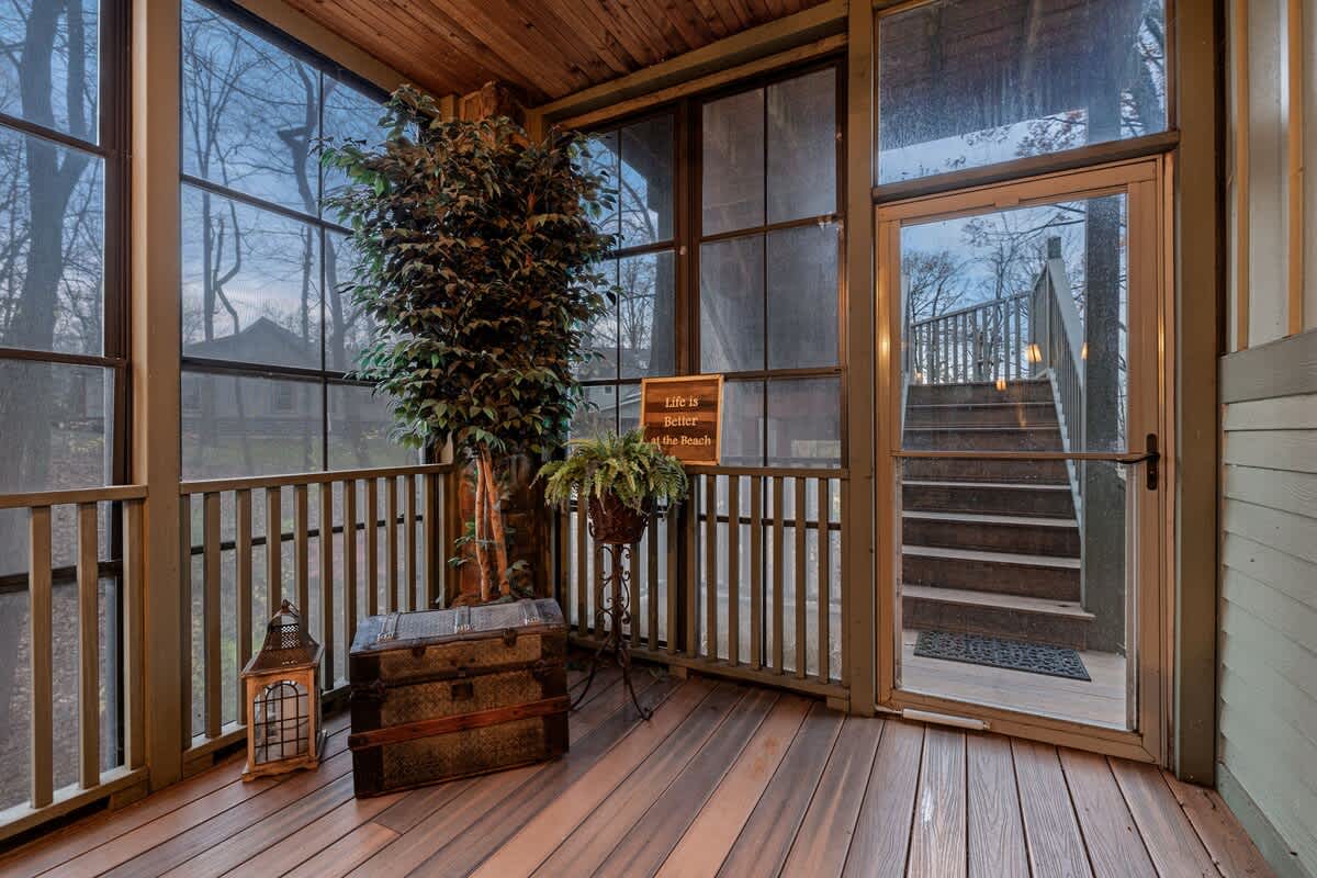 A cozy screened porch features a wooden slatted ceiling and floor, with large windows looking out to bare trees. A decorative trunk, a potted plant, and a lantern add rustic charm, while a sign reads "life is better at the beach."