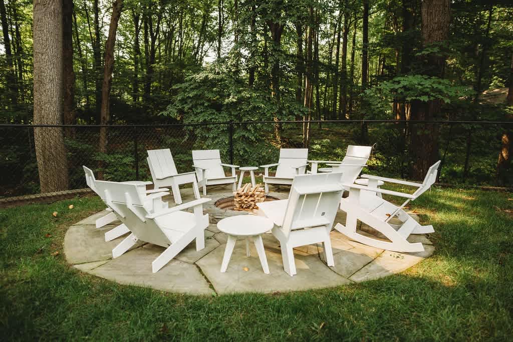 A circular stone patio hosts a fire pit surrounded by several white Adirondack chairs and small side tables, nestled in a grassy area at the edge of a dense forest.