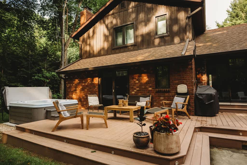 The back of a brick and wood-sided house features a multi-level wooden deck furnished with outdoor seating and a hot tub, with a BBQ grill and potted plants, all surrounded by dense green trees.