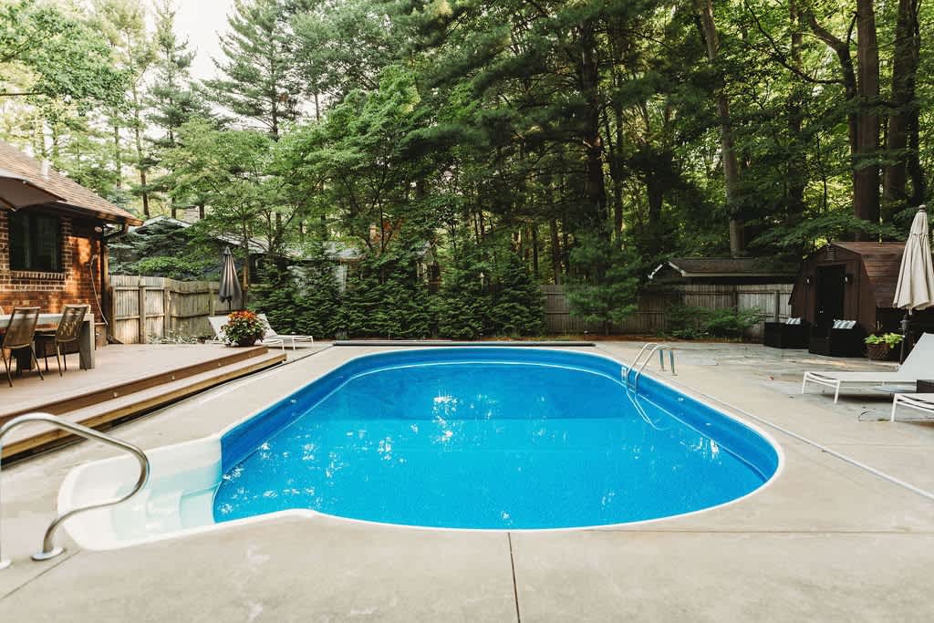 Kidney-shaped swimming pool with a concrete surround, featuring steps and handrails, set in a spacious backyard surrounded by lush green trees.