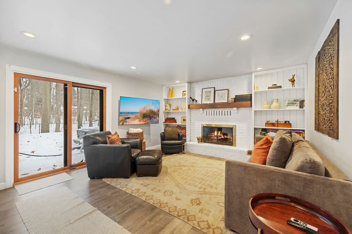 This inviting living room features a white brick fireplace with a wooden mantel, flanked by built-in shelves, a comfortable sofa, and leather armchairs, with a sliding glass door offering a view of a snowy outdoor landscape.