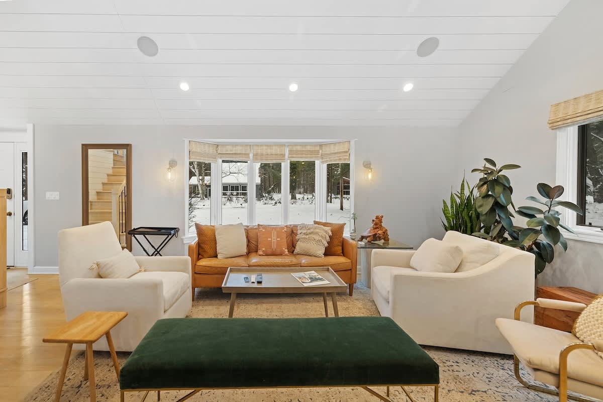 Bright living room with a shiplap ceiling, a warm orange leather sofa, two cream armchairs, and a dark green ottoman, centered around a bay window overlooking a snowy landscape.