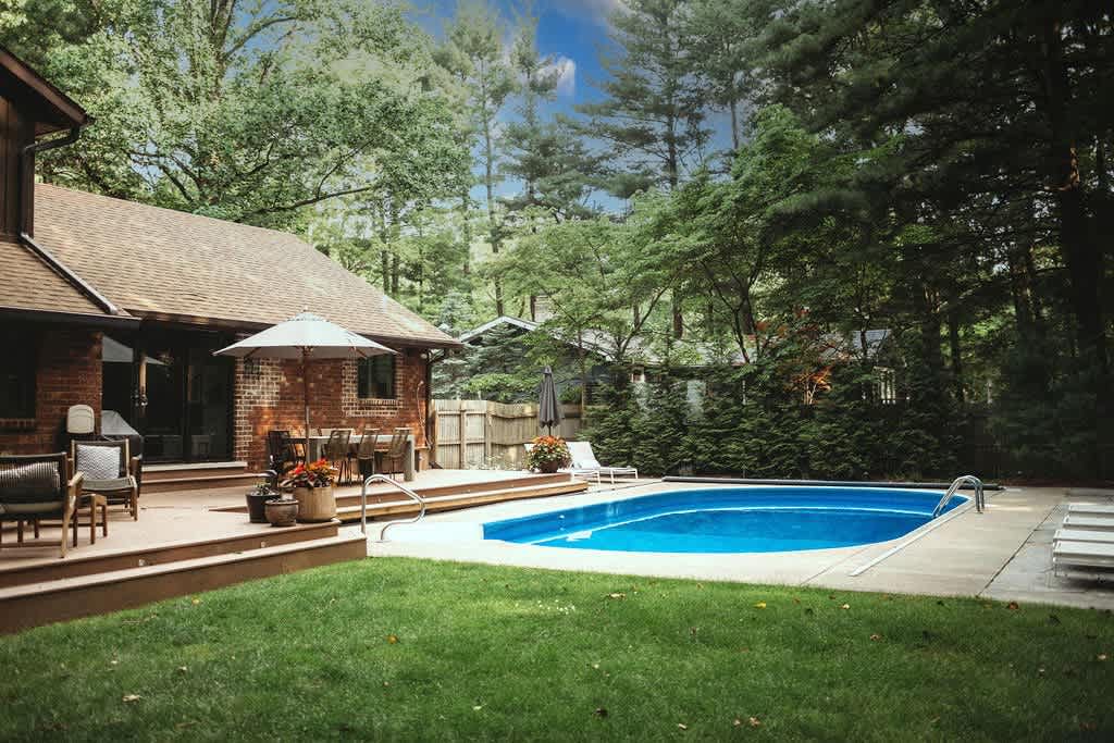 The back of the house, featuring brick and wood siding, overlooks a sparkling blue swimming pool surrounded by a concrete deck and a well-maintained green lawn, all set against a backdrop of mature trees.