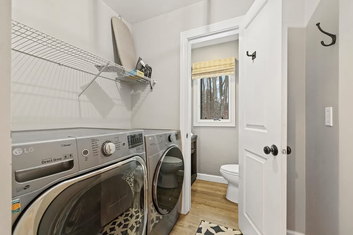 Bright laundry room with light wood-look flooring, featuring a silver LG washer and dryer, and a wire shelf above. An open door reveals a toilet and window in an adjacent bathroom.
