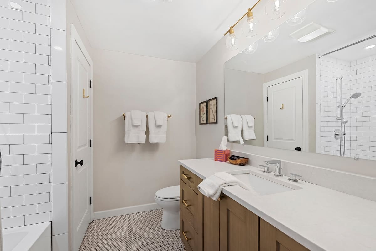 Bright bathroom with a light wood vanity, white countertop, and single sink, complemented by honeycomb floor tiles. A toilet is visible next to the vanity, and a large mirror reflects the room.