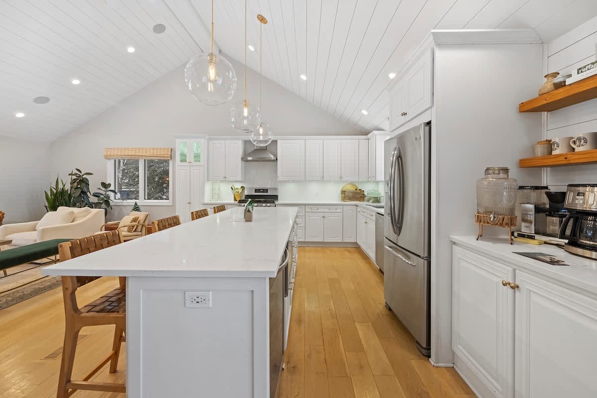 Expansive white kitchen with a long island, stainless steel refrigerator and range, and open wooden shelving, featuring a vaulted shiplap ceiling and hardwood floors, connecting to the living area.