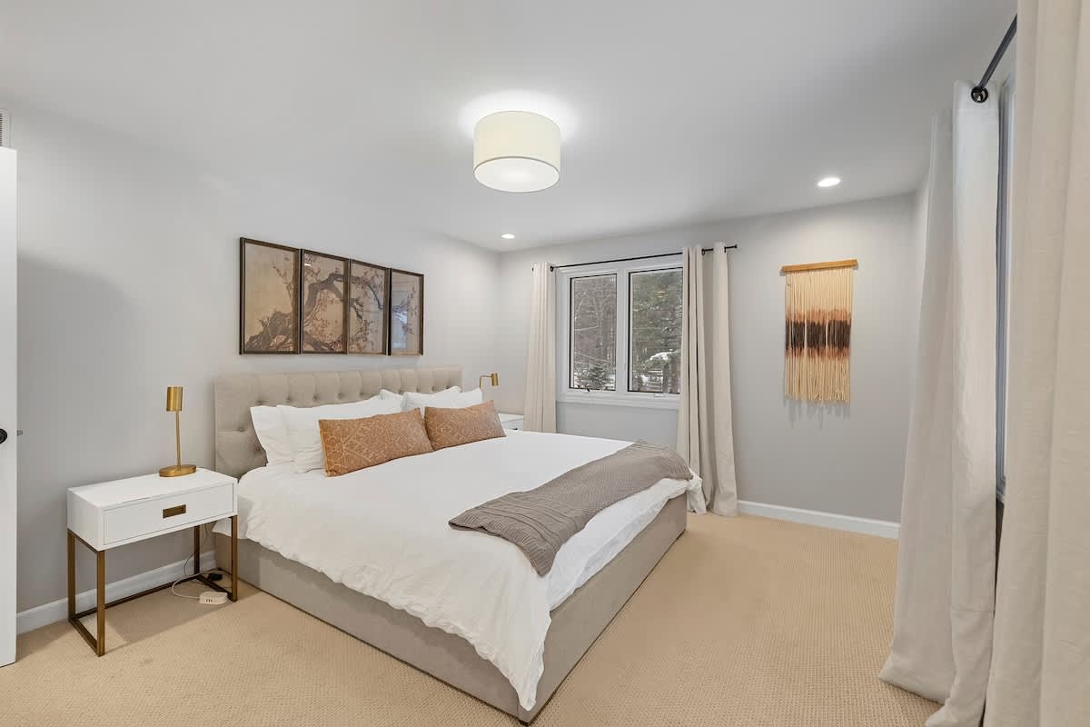 Serene bedroom featuring a queen-sized bed with a tufted headboard, white bedding, and accent pillows, flanked by modern white nightstands with gold legs. Large windows offer views of a snowy outdoor landscape, and the room has light carpet flooring.